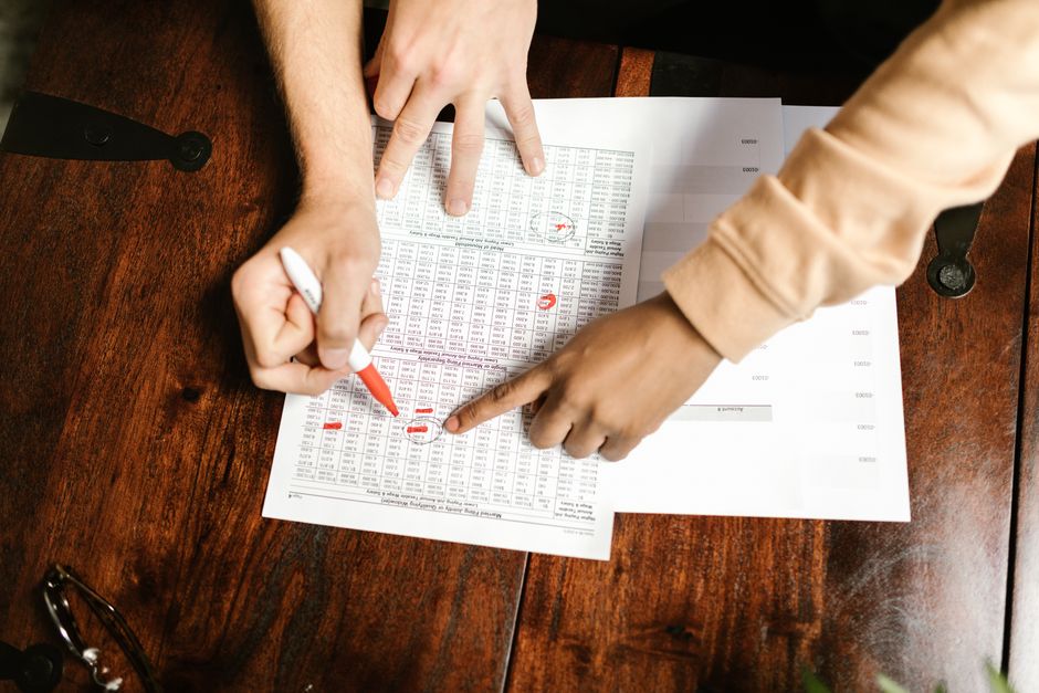 Two hands reviewing and marking financial documents with a red pen on a wooden table, top view.