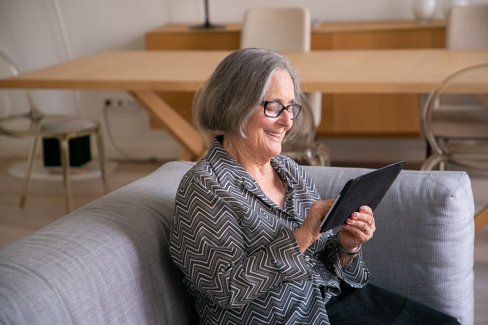 femme âgée portant des lunettes, assise sur un canapé, qui consulte une tablette